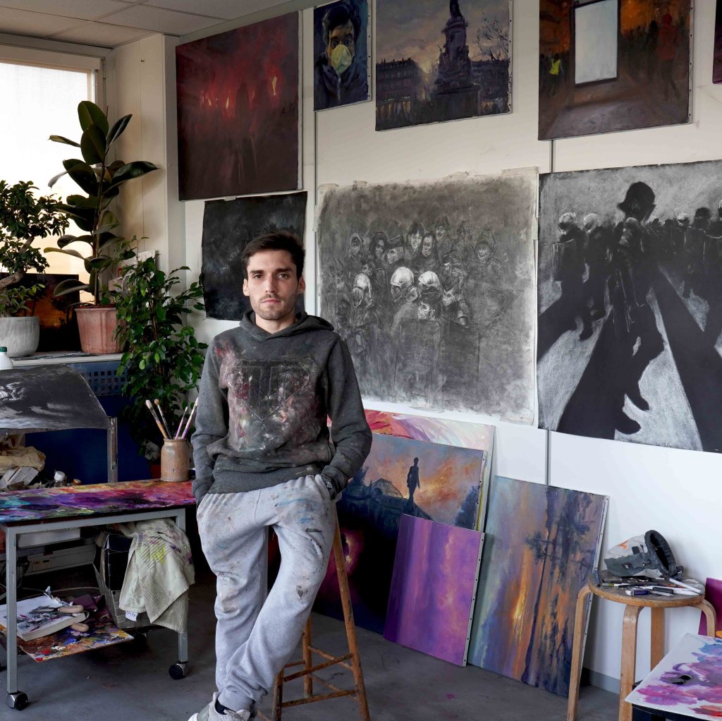 Mathieu Clément debout dans son atelier, devant de grandes peintures et dessins consacrés aux manifestations et mouvements sociaux.
